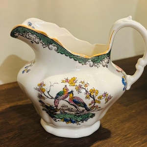 May include: A white ceramic creamer with a scalloped edge and a curved handle. The creamer has a colourful illustration of two birds on a branch with flowers. The rim is trimmed with green and gold detailing.