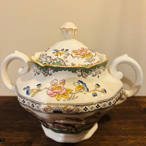 May include: A white ceramic sugar bowl with a lid, decorated with a floral pattern in pink, yellow, and blue. The bowl has two handles and a decorative rim. The lid is topped with a small finial. The design is ornate and vintage.
