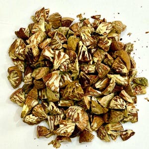 May include: A pile of chopped betel nuts, showcasing a mix of brown and cream colors. The nuts have a textured appearance with a marbled pattern. The image is taken on a white surface, highlighting the natural tones of the betel nuts.