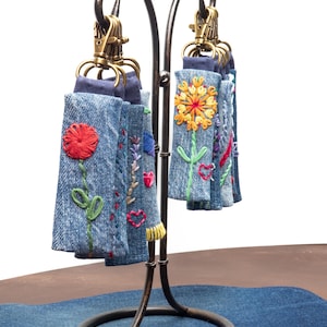 Könnte beinhalten: Schlüsselanhänger aus Jeansstoff mit gestickten Blumenmustern, präsentiert auf einem dekorativen Metallständer. Die Schlüsselanhänger weisen verschiedene farbenfrohe Blumenmuster auf, darunter eine rote Rose, eine gelbe Sonnenblume und andere florale Motive. Der Ständer hat eine dunkelbraune Oberfläche.
