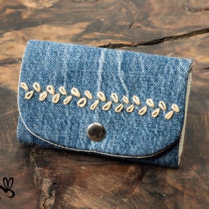 Op de afbeelding: Een kleine denim portemonnee met een klepsluiting en een zilveren drukknop. Het spijkerstofmateriaal is versierd met een rij geborduurde, beige bladachtige ontwerpen. De portemonnee ligt op een donkere houten achtergrond.