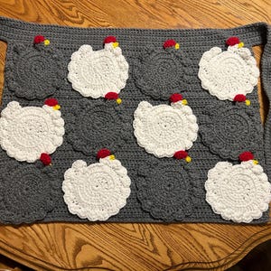 May include: A handmade gray crochet apron featuring a repeating pattern of white crochet chickens with red combs and yellow beaks. The apron has a pocket design and long ties for securing around the waist.