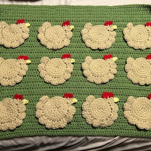 May include: A handmade, crocheted blanket featuring a repeating pattern of tan-colored chickens with red combs and yellow beaks on a green background. The blanket is made of yarn and has a textured appearance, perfect for home decor.