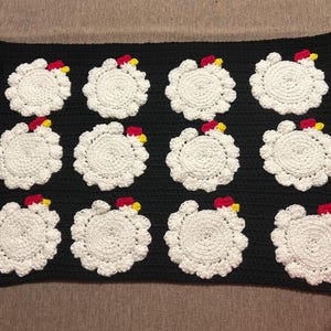 May include: Black crochet apron featuring twelve white crochet chicken appliques with red and yellow accents. The apron has long black ties for securing around the waist.