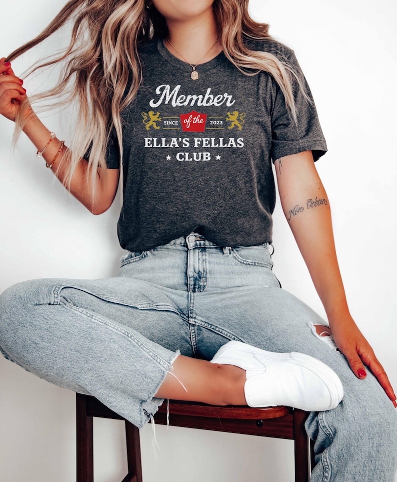 May include: Dark gray t-shirt with the text "Member of the Ella's Fellas Club" in white, red, and gold. The shirt features a decorative design with the year 2023. The person is wearing light blue jeans and white shoes.
