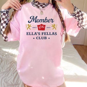 May include: A light pink t-shirt with the text "Member of the Ella's Fellas Club" in navy blue and gold. The shirt features a decorative design with two lions and the year 2023.