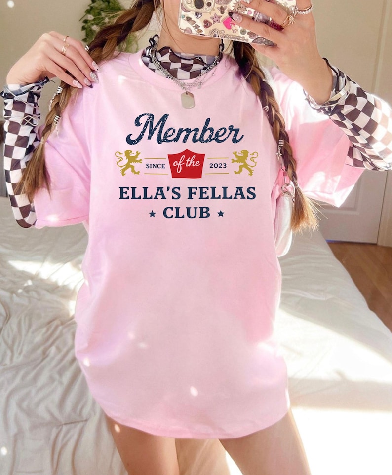 May include: A light pink t-shirt with the text "Member of the Ella's Fellas Club" in navy blue and gold. The shirt features a decorative design with two lions and the year 2023.