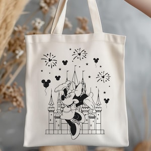 K&ouml;nnte beinhalten: Cremefarbene Tragetasche mit einer Schwarz-Wei&szlig;-Illustration eines Schlosses, Feuerwerks und Minnie Mouse. Die Tasche hat ein schlichtes Design mit zwei Henkeln und wird von einer Person gehalten.