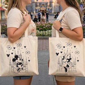 K&ouml;nnte beinhalten: Zwei cremefarbene Tragetaschen mit schwarzen Illustrationen von Mickey und Minnie Mouse vor einem Schloss, mit Feuerwerk und Herzen. Die Taschen werden von zwei Personen vor einem Schloss gehalten.