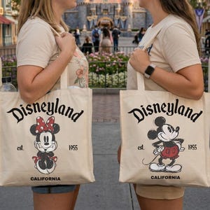 May include: Two beige tote bags with the Disneyland logo, Minnie Mouse and Mickey Mouse illustrations. The bags read "Disneyland est. 1955 CALIFORNIA". The background features a castle and buildings.