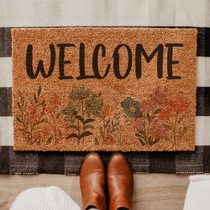 May include: A rectangular tan coir doormat featuring the word WELCOME in black capital letters, decorated with a colorful floral print along the bottom edge, layered over a black and white buffalo check fabric rug on a light wood floor.