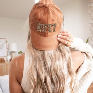 May include: A brown baseball cap with the word "WIFEY" in leopard print across the front. The cap is made of a soft material and has a curved brim. The hat is a casual accessory.