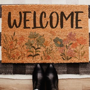 May include: A rectangular tan coir doormat featuring the word WELCOME in bold black lettering. The lower portion of the mat displays a printed botanical design with various wildflowers in shades of green, orange, pink, and muted red.