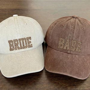 May include: Two baseball caps, one beige and one brown. The beige cap has the word "BRIDE" in a leopard print font. The brown cap has the word "BABE" in a leopard print font. Both caps have a curved brim.
