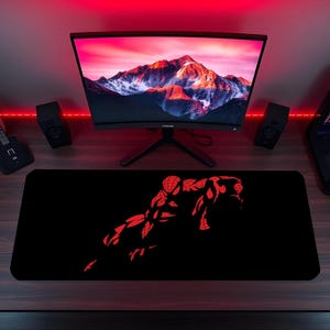 May include: A rectangular black desk mat featuring a red graphic illustration of a superhero in a crouching pose. The mat is positioned on a dark wood desk surface in front of a computer monitor displaying a mountain landscape and two black speakers.
