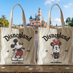 May include: Two beige canvas tote bags with black text that reads "Disneyland" above cartoon illustrations of Mickey and Minnie Mouse. The bags also have "est. 1955" and "CALIFORNIA" printed on them. The background shows a castle.