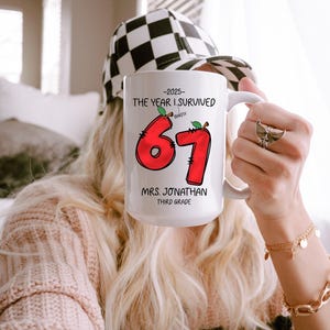 May include: White ceramic mug with the text "-2025- THE YEAR I SURVIVED BARELY 67 MRS. JONATHAN THIRD GRADE" in black and red. The number 67 is designed with a red apple theme. The mug is held by a person wearing a black and white checkered hat.
