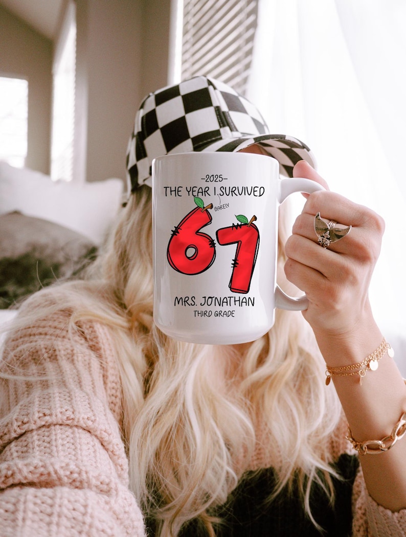 May include: White ceramic mug with the text "-2025- THE YEAR I SURVIVED BARELY 67 MRS. JONATHAN THIRD GRADE" in black and red. The number 67 is designed with a red apple theme. The mug is held by a person wearing a black and white checkered hat.