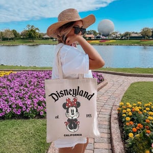 May include: A beige canvas tote bag with the Disneyland logo and a Minnie Mouse graphic. The bag features the text "est. 1955 CALIFORNIA". The bag is carried outdoors near a lake and flower beds.