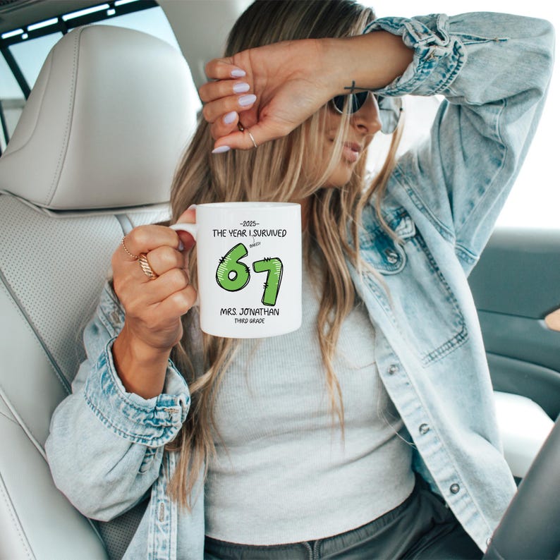May include: White ceramic mug with the text "-2025- THE YEAR I SURVIVED 67 MRS JONATHAN THIRD GRADE" in black and green. The mug is held by a person wearing a denim jacket.