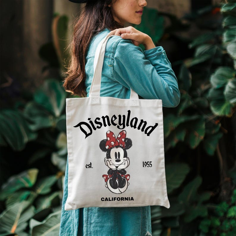 May include: White canvas tote bag featuring the Disneyland logo in black script, with a Minnie Mouse graphic wearing a red bow. The text "est. 1955 CALIFORNIA" is also printed on the bag. The bag has two white handles.