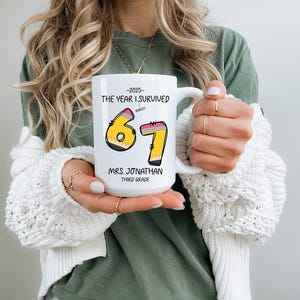 May include: White ceramic mug with a handle, featuring the text "-2025 THE YEAR I SURVIVED" above the numbers "67" designed as pencils. Below the numbers is "MRS. JONATHAN THIRD GRADE".