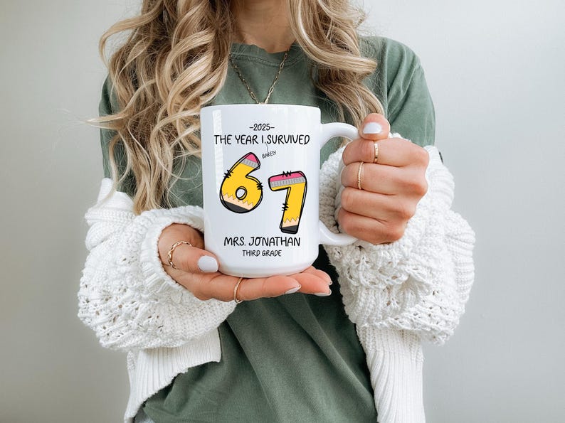 May include: White ceramic mug with a handle, featuring the text "-2025 THE YEAR I SURVIVED" above the numbers "67" designed as pencils. Below the numbers is "MRS. JONATHAN THIRD GRADE".