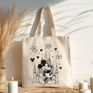 K&ouml;nnte beinhalten: Cremefarbene Tragetasche mit einer schwarz-wei&szlig;en Illustration von Mickey Mouse vor einem Schloss, mit Feuerwerk und Mickey Mouse-Kopf-Ballons. Die Tasche hat zwei Henkel und ist aus einem robusten Stoff gefertigt.