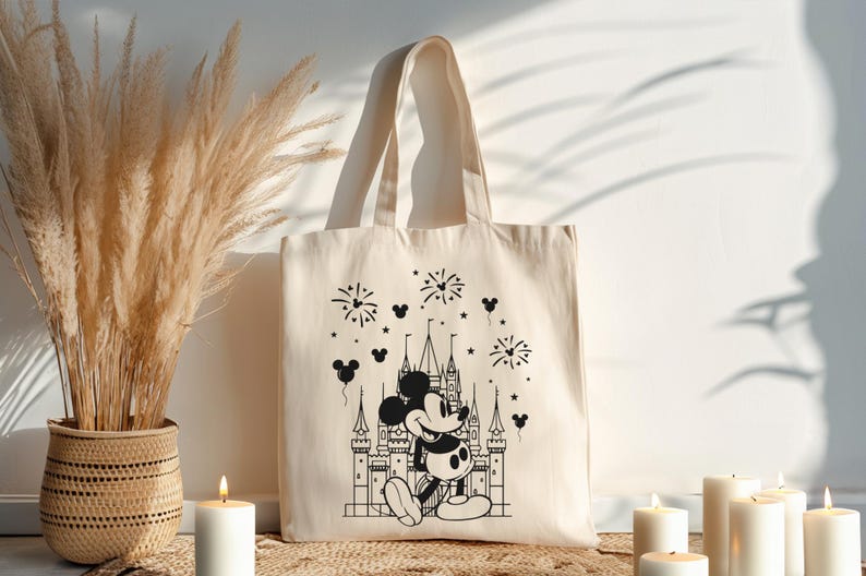 K&ouml;nnte beinhalten: Cremefarbene Tragetasche mit einer schwarz-wei&szlig;en Illustration von Mickey Mouse vor einem Schloss, mit Feuerwerk und Mickey Mouse-Kopf-Ballons. Die Tasche hat zwei Henkel und ist aus einem robusten Stoff gefertigt.