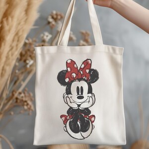 K&ouml;nnte beinhalten: Cremefarbene Tragetasche mit einer Schwarz-Wei&szlig;-Illustration von Minnie Mouse. Minnie ist mit ihrer charakteristischen rot-wei&szlig; gepunkteten Schleife und passenden Akzenten dargestellt. Die Tasche hat zwei Henkel und wird von einer Person gehalten.
