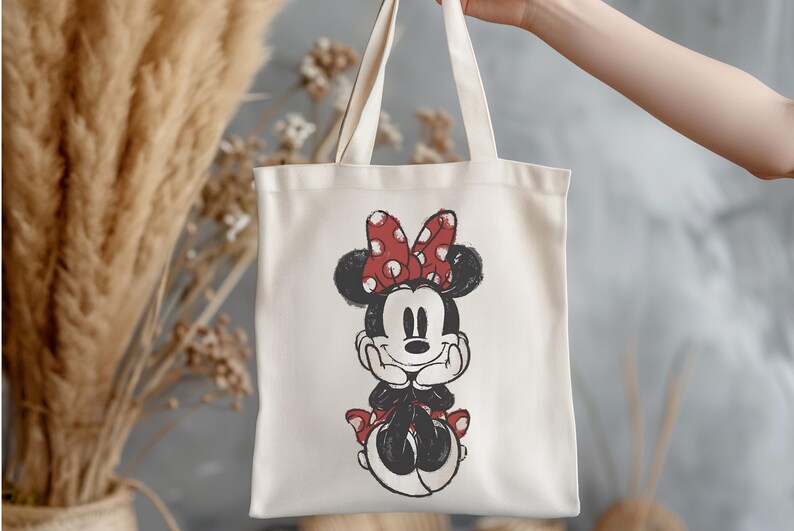 K&ouml;nnte beinhalten: Cremefarbene Tragetasche mit einer Schwarz-Wei&szlig;-Illustration von Minnie Mouse. Minnie ist mit ihrer charakteristischen rot-wei&szlig; gepunkteten Schleife und passenden Akzenten dargestellt. Die Tasche hat zwei Henkel und wird von einer Person gehalten.