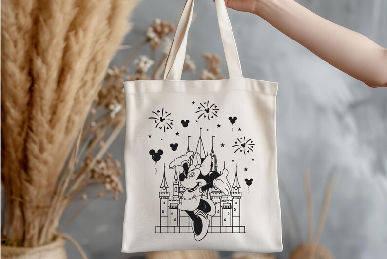 K&ouml;nnte beinhalten: Cremefarbene Tragetasche mit einer Schwarz-Wei&szlig;-Illustration eines Schlosses, Feuerwerks und Minnie Mouse. Die Tasche hat ein schlichtes Design mit zwei Henkeln und wird von einer Person gehalten.