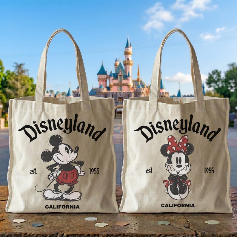 May include: Two beige canvas tote bags with black text that reads "Disneyland" above cartoon illustrations of Mickey and Minnie Mouse. The bags also have "est. 1955" and "CALIFORNIA" printed on them. The background shows a castle.