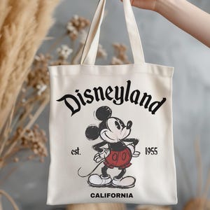 May include: Cream-colored tote bag featuring a vintage-style Mickey Mouse graphic. The bag displays the text "Disneyland" in a stylized font, along with "est. 1955" and "CALIFORNIA". The iconic cartoon character is depicted in a classic pose.
