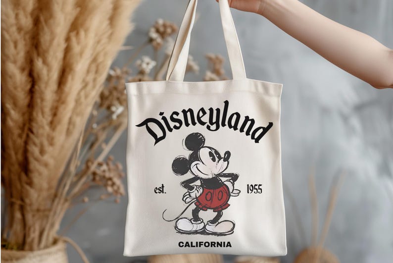 May include: Cream-colored tote bag featuring a vintage-style Mickey Mouse graphic. The bag displays the text "Disneyland" in a stylized font, along with "est. 1955" and "CALIFORNIA". The iconic cartoon character is depicted in a classic pose.