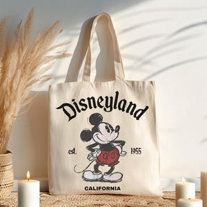 May include: Cream-colored tote bag featuring the Disneyland logo and a vintage Mickey Mouse illustration. The bag includes the text "est. 1955" and "CALIFORNIA". The bag has two cream-colored handles.