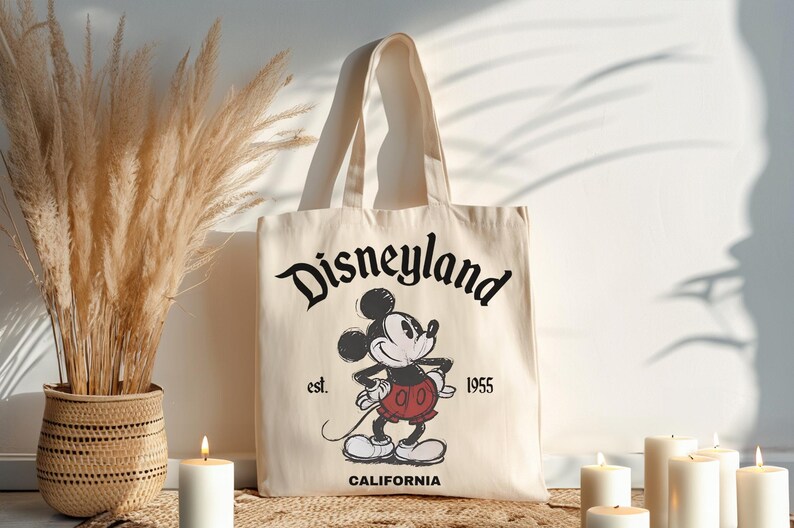 May include: Cream-colored tote bag featuring the Disneyland logo and a vintage Mickey Mouse illustration. The bag includes the text "est. 1955" and "CALIFORNIA". The bag has two cream-colored handles.