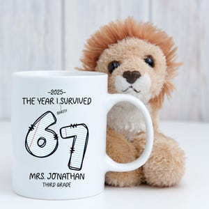 May include: White ceramic mug with black text that reads "-2025- THE YEAR I SURVIVED BARELY 67 MRS. JONATHAN THIRD GRADE." A plush lion toy is behind the mug. The mug has a white handle.