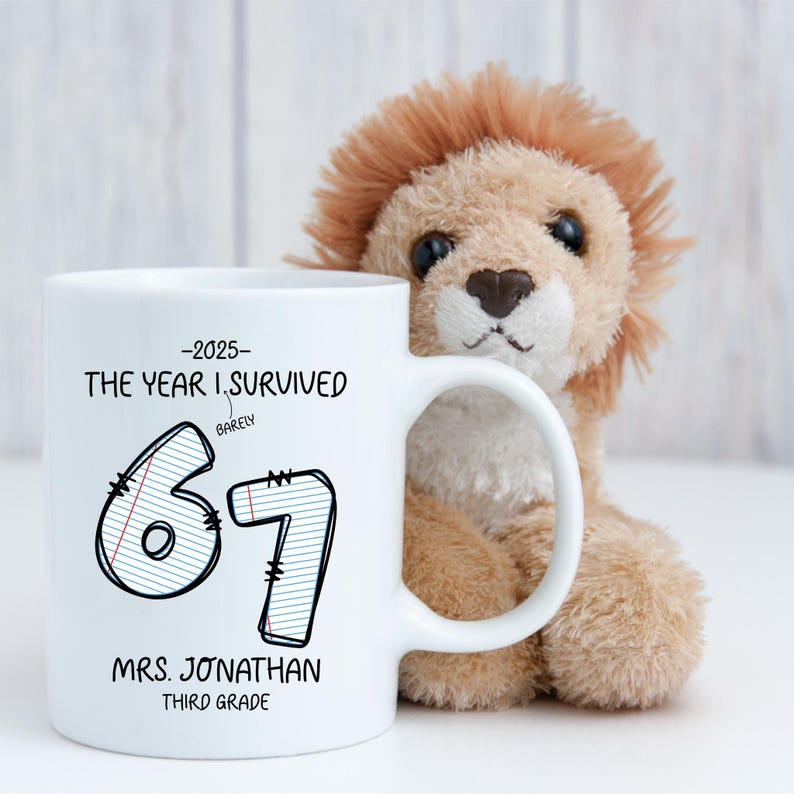 May include: White ceramic mug with black text that reads "-2025- THE YEAR I SURVIVED BARELY 67 MRS. JONATHAN THIRD GRADE." A plush lion toy is behind the mug. The mug has a white handle.