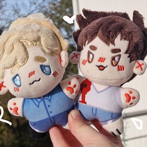 Puede incluir: Dos muñecos de peluche con rasgos de dibujos animados se sostienen en una mano. Uno tiene pelo rubio y un atuendo azul, el otro tiene pelo castaño y un atuendo blanco y azul. Ambos muñecos tienen detalles faciales bordados y pies con forma de pata.