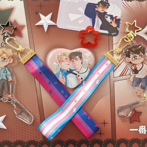 Könnte beinhalten: Sammlung von Anime-Schlüsselanhängern und Accessoires. Enthält zwei Schlüsselanhänger mit Cartoon-Charakteren, eine herzförmige Anstecknadel und zwei Schlüsselbänder mit Designs der bisexuellen und transgender Pride-Flagge. Dekorative Sterne und Comic-Hintergrund.