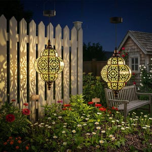 May include: Two ornate, hanging solar lanterns with intricate cut-out patterns illuminate a garden at dusk. The lanterns are suspended from black cords, casting decorative light patterns on a white picket fence and surrounding foliage.