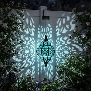 May include: A hanging teal-colored lantern with an intricate, openwork design, casting a patterned light on a white fence. The lantern is suspended by a black cord and surrounded by lush greenery and flowers, creating a serene outdoor ambiance.