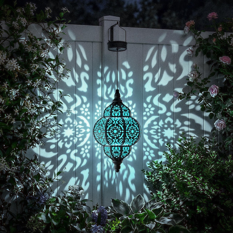 May include: A hanging teal-colored lantern with an intricate, openwork design, casting a patterned light on a white fence. The lantern is suspended by a black cord and surrounded by lush greenery and flowers, creating a serene outdoor ambiance.