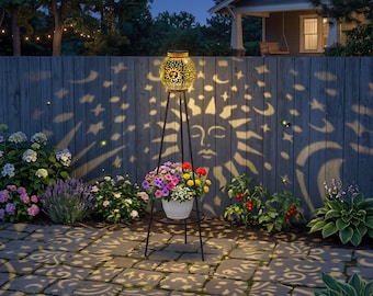 May include: A tall, black metal tripod plant stand featuring a spherical, gold-toned solar lantern at the top. The lantern casts intricate, glowing projections of suns, moons, and stars onto the surrounding fence and stone patio surface.