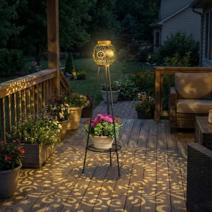 May include: A black metal outdoor solar lantern with a decorative cut-out design, casting intricate patterns of light on a wooden deck. The lantern is supported by a tripod stand and has a small shelf holding a potted plant.