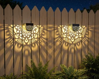 May include: Two black solar-powered lights mounted on a wooden fence, casting a decorative pattern of light. The lights are positioned against a night sky backdrop, with green plants in the foreground. The lights have a decorative floral design.