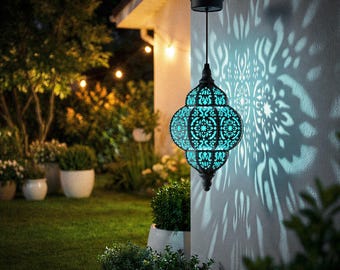 Puede incluir: Una linterna colgante turquesa con intrincados patrones calados, proyectando un diseño de luz decorativo sobre una pared blanca. La linterna está suspendida de un gancho negro y está rodeada de vegetación e iluminación exterior, creando un ambiente cálido.