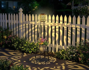 May include: A solar-powered outdoor garden light with a decorative patterned shade, casting intricate shadows on a white picket fence and surrounding foliage. The light is supported by a black metal tripod stand, with a small planter of flowers below.