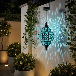 May include: A black and turquoise solar-powered lantern with an intricate, openwork design hangs from a hook. The lantern casts a decorative light pattern on the wall. Green ivy and small potted plants with fairy lights surround the lantern.